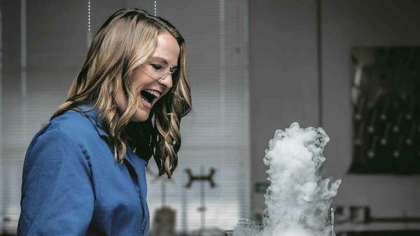 Kate Biberdorf is pictured smiling in a laboratory. A white cloud rises from a beaker. Kate is wearing a lab coat and protective glasses.