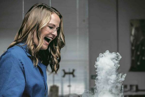 Kate Biberdorf is pictured smiling in a laboratory. A white cloud rises from a beaker. Kate is wearing a lab coat and protective glasses.